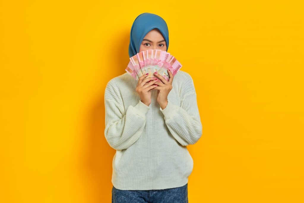 how to become millionier in 20 age - Beautiful Asian Muslim woman in white sweater covering face with cash money in Indonesian rupiah banknotes isolated over yellow background. ​People religious lifestyle concept