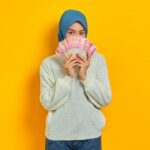 how to become millionier in 20 age - Beautiful Asian Muslim woman in white sweater covering face with cash money in Indonesian rupiah banknotes isolated over yellow background. ​People religious lifestyle concept