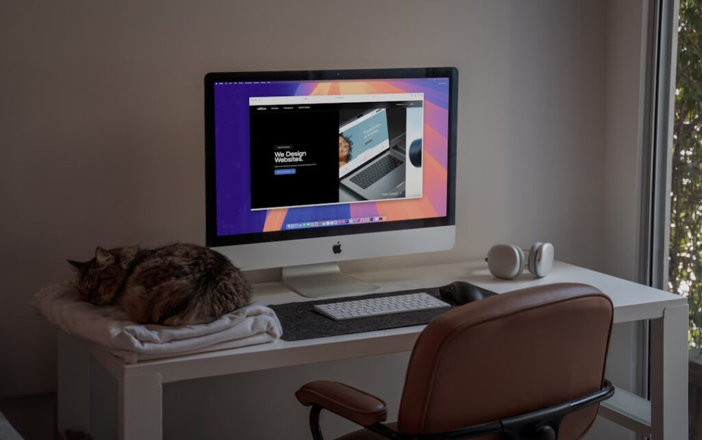 remote work productivity - A peaceful home office setup featuring an iMac showcasing a modern web design project, with a comfortable brown leather chair, wireless headphones, and a sleeping cat on a soft blanket. The natural light filtering through the window adds a calming atmosphere, making this an ideal space for creativity and productivity. Perfect inspiration for remote workers, designers, or anyone who loves minimalist workspaces with a touch of coziness.