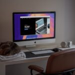 remote work productivity - A peaceful home office setup featuring an iMac showcasing a modern web design project, with a comfortable brown leather chair, wireless headphones, and a sleeping cat on a soft blanket. The natural light filtering through the window adds a calming atmosphere, making this an ideal space for creativity and productivity. Perfect inspiration for remote workers, designers, or anyone who loves minimalist workspaces with a touch of coziness.
